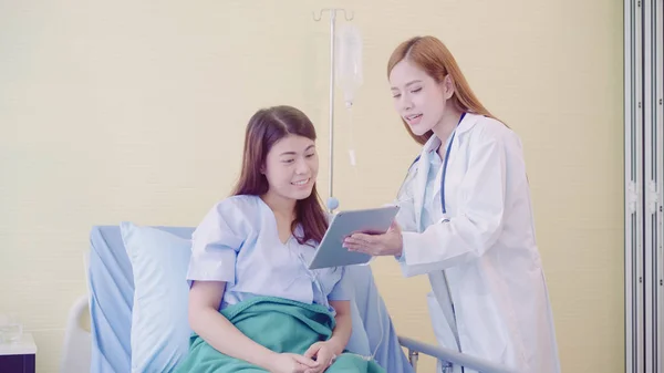 Belo médico asiático inteligente e paciente discutindo e explicando algo com tablet em mãos de médico enquanto fica na cama do paciente no hospital. Conceito de medicina e cuidados de saúde . — Fotografia de Stock