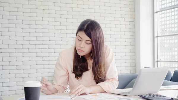 Vakker, ung, smilende asiatisk kvinne som jobber på pc-en i stua hjemme. En asiatisk forretningskvinne som skriver notatbok, finans og kalkulator på hjemmekontoret. Nyter tid hjemme-konseptet . – stockfoto