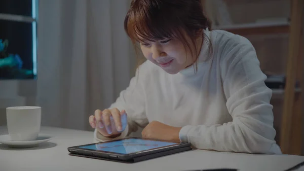Ung asiatisk kvinne som jobber sent med nettbrett, sjekker sosiale medier mens hun slapper av på skrivebordet om natten i stua hjemme. Nyter tid hjemme-konseptet . – stockfoto
