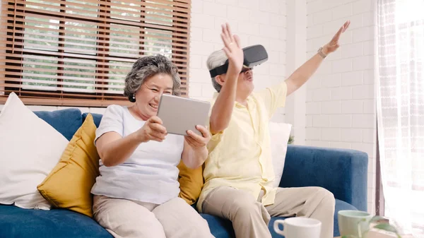 Asian elderly couple using tablet and virtual reality simulator playing games in living room, couple feeling happy using time together lying on sofa at home. Lifestyle Senior family at home concept. Stock Picture