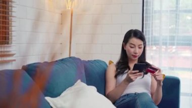 Genç gülümseyen Asyalı kadın evde oturma odasında dinlenirken kredi kartıyla online alışveriş yapıyor. Ev kavramında yaşam tarzı kadınları.