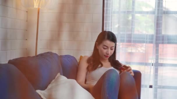 年轻微笑的亚洲女人在家里客厅里躺在沙发上 用平板电脑用信用卡在网上购物 居家妇女的生活方式概念 — 图库视频影像