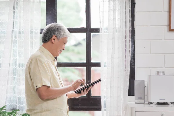 Asiatiska äldre man använder Tablet PC kontrollera sociala medier nära fönster i vardagsrum hemma. Livsstil Senior män hemma Concept. — Stockfoto