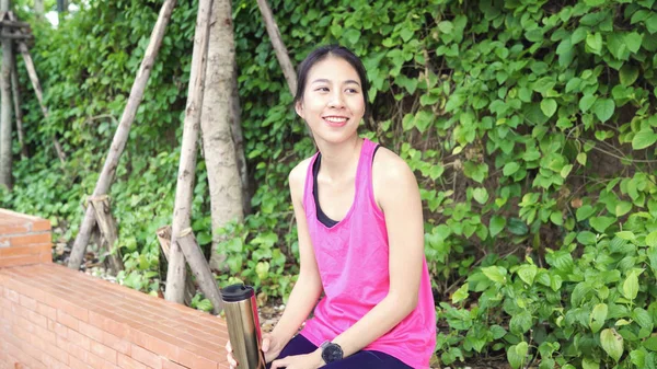 Gesunde schöne junge asiatische Läuferin trinkt Wasser, weil sie sich nach dem Laufen auf der Straße im städtischen Stadtpark müde fühlt. — Stockfoto