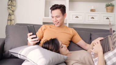 Asya çift Smartphone ve dizüstü evde oturma odasında sosyal medya denetimi kullanarak, tatlı çift evde rahat zaman kanepede yatarken aşk anın tadını çıkarın. Yaşam tarzı çift evde kavramı relax.