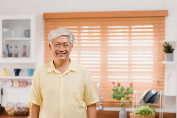 Aasialainen iäkäs mies hymyilee onnellisena ja etsii kameraa rentoutuessaan keittiössä kotona. Elämäntapa vanhemmat miehet kotona käsite . — kuvapankkivalokuva