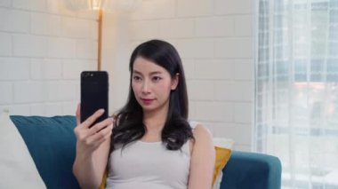 Genç Asyalı kadın akıllı telefon kullanarak sosyal medyayı kontrol ediyor. Evdeki oturma odasında dinlenirken kanepede yatarken gülümsüyor ve mutlu oluyor. Ev kavramında yaşam tarzı kadınları.