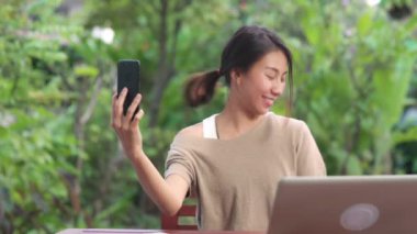 Sosyal medyada cep telefonuyla selfie çeken Asyalı bir kadın. Kadın rahat rahat bahçede sabah masada oturan alışveriş torbalarını gösteriyor. Yaşam tarzı kadınları ev kavramında rahatlar.