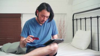 Genç Asyalı adam tablet ve kredi kartı ekommersi kullanıyor, erkek rahat rahat evde yatak odasında yatakta otururken online alışveriş yapıyor. Gençler internetten alışveriş yapıyor, online iletişim konsepti..