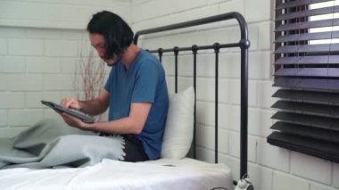 Genç Asyalı adam tablet kullanıyor. Sosyal medyayı kontrol ediyor. Mutlu bir şekilde gülümsüyor. Sabah uyandıktan sonra yatakta yatıyor..