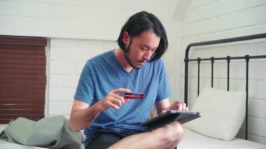 Genç Asyalı adam tablet ve kredi kartı ekommersi kullanıyor, erkek rahat rahat evde yatak odasında yatakta otururken online alışveriş yapıyor. Gençler internetten alışveriş yapıyor, online iletişim konsepti..