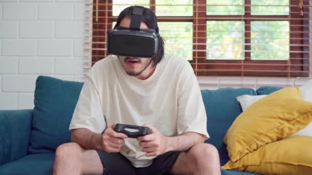 亚洲男人在客厅里用眼镜 虚拟现实模拟器和操纵杆玩电子游戏 男人在家里躺在沙发上放松一下 感觉很快乐 男人在家里玩游戏放松 — 图库视频影像