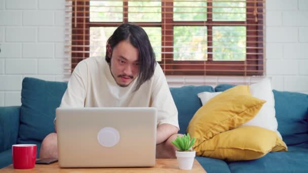 自宅で働くフリーランスのアジア人男性 リビングルームのソファの上のラップトップ上の男性の創造的 ビジネス若い男の所有者起業家 コンピュータを再生 現代の家のコンセプトで職場でソーシャルメディアをチェック — ストック動画