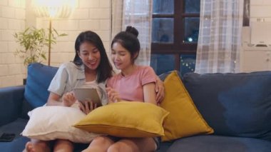 Evde tablet kullanan lezbiyen lgbt kadın çift, teknolojiyi kullanarak mutlu olan Asyalı kadın gece oturma odasında kanepeydeyken internette sosyal medyayı birlikte kontrol ediyor. Lover tatil konsepti kutlamak.