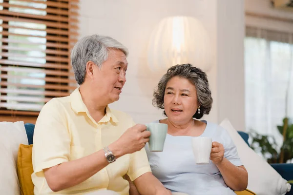 Asiatisches älteres Paar, das warmen Kaffee trinkt und zu Hause im Wohnzimmer miteinander spricht, genießen Liebesmomente, während sie zu Hause entspannt auf dem Sofa liegen. Lifestyle Seniorenfamilie zu Hause Konzept. — Stockfoto