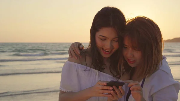 Młoda Azjatycka Para lesbijek za pomocą inteligentnego telefonu w pobliżu plaży. Piękne kobiety LGBT para szczęśliwy relaks cieszyć się chwila miłości, gdy zachód słońca wieczorem. Styl życia lesbijek para podróży na plaży koncepcji — Zdjęcie stockowe