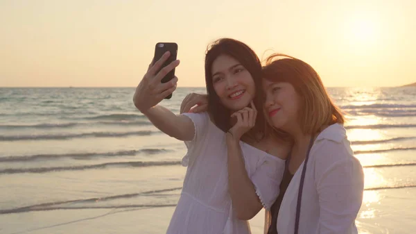 Unge asiatiske lesbiske par som bruker smarttelefon ved stranden. Vakre kvinner lgbt parret glade kjærlighets- øyeblikk når solnedgang om kvelden. Livsstilslesbiske parreiser på strandkonseptet – stockfoto