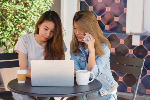 Mujeres jóvenes asiáticas que trabajan en el ordenador portátil usando y mirando teléfono inteligente y beber café mientras están sentadas en la cafetería. Estilo de vida de las mujeres de comunicación y trabajo en concepto de cafetería . —  Fotos de Stock