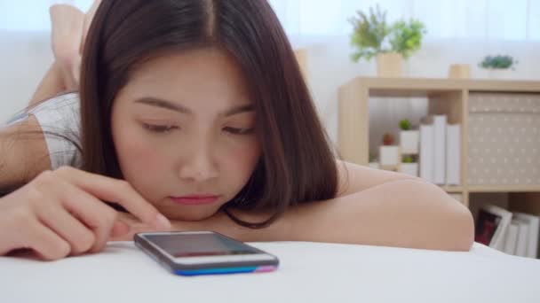 朝起きてベッドに横たわりながら幸せな笑顔をソーシャルメディアチェックをスマホでチェックする若いアジア人女性 自宅の寝室でくつろぐ美しい魅力的な日本の女の子 — ストック動画