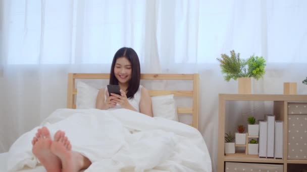 スローモーション 朝起きた後にベッドに横たわっている間に幸せな笑顔を感じるソーシャルメディアをチェックするスマートフォンを使用する若いアジアの女性 自宅の寝室でリラックスして笑顔の魅力的な日本の女の子 — ストック動画