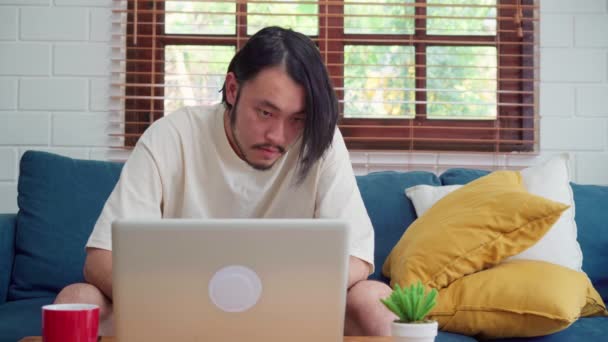 自宅で働くフリーランスのアジア人男性 リビングルームでソファのコーヒーを飲む上でラップトップ上の男性の創造的 ビジネス若い男の所有者起業家 現代の家の概念で職場でコンピュータを再生します — ストック動画