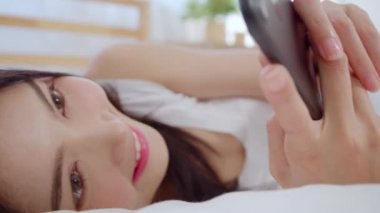 Akıllı telefon kontrol sosyal medya kontrol kullanarak genç Asyalı kadın sabah uyandıktan sonra yatakta yatarken mutlu gülümseyerek duygu, Güzel çekici Japon kız evde yatak odasında dinlenmek gülümseyen.