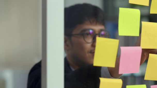 アジアのクリエイティブな男は ガラス板に付箋を貼ります 若いプロのビジネス男性は ロフトオフィスのコンセプトで情報 紙のリマインダー ビジネス状況 スタートアップを書きます スローモーション — ストック動画
