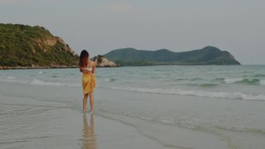 Kum sahilinde yürüyen Asyalı bir kadın. Bikini genç mutlu kadın dinlenmek ve eğlenceli tropik deniz yakınında yürürken gün batımı sırasında tatil, tatil, yaz gezisi kavramı. Yavaş çekim çekimi.