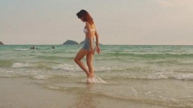 Kum sahilinde yürüyen Asyalı bir kadın. Bikini genç mutlu kadın dinlenmek ve eğlenceli tropik deniz yakınında yürürken gün batımı sırasında tatil, tatil, yaz gezisi kavramı. Yavaş çekim çekimi.