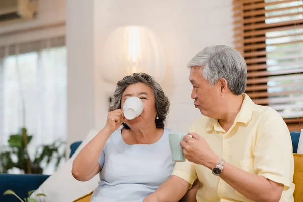 温かいコーヒーを飲み、自宅のリビングルームで一緒に話すアジアの高齢夫婦は、自宅でリラックスした時にソファに横たわりながらラブモーメントを楽しむ。在宅ライフスタイルシニアファミリーコンセプト. — ストック写真