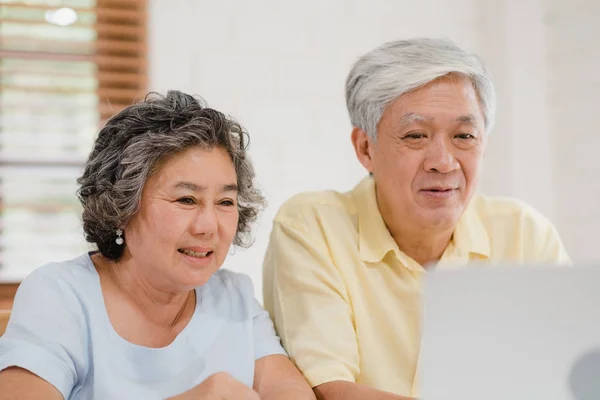 Asian elderly couple using laptop conference with doctor about medicine information in living room, couple using time together while lying on sofa at home. Senior family health at home concept. — Stock Photo, Image