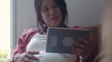 Hamilelik bilgileri için tablet kullanan Asyalı hamile bir kadın. Anne kendini mutlu hissederken pozitif ve huzurlu gülümserken çocuğuyla ilgilenirken oturma odasında kanepede uzanmış ev konseptinde..