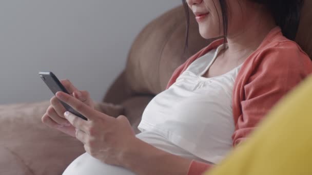 携帯電話検索の妊娠情報を使用して若いアジアの妊婦 母は幸せな笑顔正と平和ながら世話をします彼女の子供は自宅のコンセプトでリビングルームでソファに横たわって — ストック動画