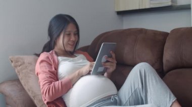 Hamilelik bilgileri için tablet kullanan Asyalı hamile bir kadın. Anne kendini mutlu hissederken pozitif ve huzurlu gülümserken çocuğuyla ilgilenirken oturma odasında kanepede uzanmış ev konseptinde..