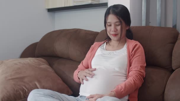 若いアジア妊娠中の女性は彼女の子供と話す彼女の腹を保持する ママ幸せな笑顔正と平和ながら世話赤ちゃんを取ります 妊娠は自宅のコンセプトでリビングルームでソファに横たわっています — ストック動画