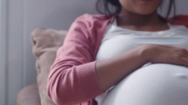 Genç Asyalı Hamile kadın karnını tutarak çocuğuyla konuşuyor. Anne mutlu gülümseyerek olumlu ve huzurlu bebek bakım almak bebek, hamilelik evde oturma odasında kanepede yatan kavram. Yavaş çekim