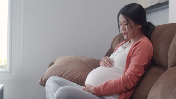 彼女の腹を抱えた若いアジアの妊婦が子供と話している 幸せな笑顔を感じる肯定的で平和なケア赤ちゃん 自宅のコンセプトでリビングルームでソファに横たわっている妊娠 スローモーション — ストック動画