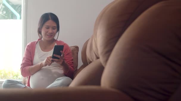 携帯電話検索妊娠情報を使用して若いアジアの妊婦 お母さんは 自宅のリビングルームでソファに横たわっている彼女の子供の世話をしながら 肯定的で平和な笑顔を幸せに感じています スローモーション — ストック動画