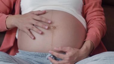Genç Asyalı hamile kadın karnını tutuyor ve çocuğuyla konuşuyor. Annem mutlu, pozitif ve huzurlu gülümserken kendine iyi bak bebeğim. Oturma odasındaki kanepede uzanmış hamilelik..