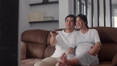 Genç Asyalı hamile çift televizyon izliyor. Anne ve baba mutlu bir şekilde gülümserken bebeğe bakıyorlar. Oturma odasındaki kanepede uzanıyorlar..
