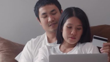 Genç Asyalı hamile çift evde online alışveriş yapıyor. Annem ve babam, ev konseptinde oturma odasındaki kanepede uzanmış bebek ürünleri alırken dizüstü bilgisayar ve kredi kartı kullanmaktan mutlu oluyorlar..