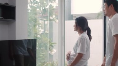 Genç Asya Hamile çift sarılmak ve çocukları ile konuşurken göbek tutarak. Anne ve baba mutlu gülümseyerek huzurlu bir şekilde bakarken bebek, evde oturma odasında pencerenin yanında hamilelik. Yavaş çekim.