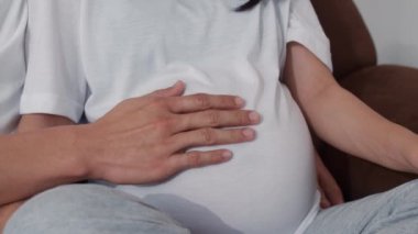 Genç Asyalı Hamile çift göbek tutan kalp işareti yapıyor. Anne ve baba mutlu mutlu gülümseyerek huzurlu bir şekilde bakarken bebek, hamilelik evde oturma odasında kanepede yatan konsept. Yavaş çekim.
