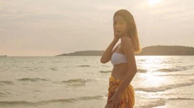 Asyalı kadın kum sahilde gülümsüyor. Bikini genç mutlu kadın dinlenmek ve tropikal deniz yakınında eğlenceli bir gülümseme zaman gün batımı sırasında tatil, tatil, yaz gezisi kavramı. Yavaş çekim çekimi.