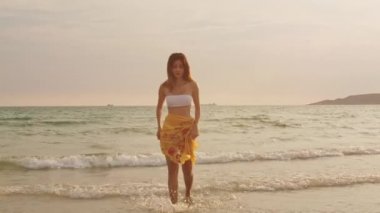 Asyalı kadın kum sahilde gülümsüyor. Bikini genç mutlu kadın dinlenmek ve tatil sırasında gün batımı tropikal deniz yakınında eğlenceli bir gülümseme; tatil; yaz gezisi konsepti. Yavaş çekim çekimi.