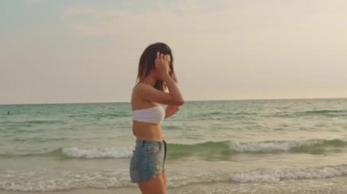 Kum sahilinde yürüyen Asyalı bir kadın. Bikini genç mutlu kadın dinlenmek ve eğlenceli tropik deniz yakınında yürürken gün batımı sırasında tatil, tatil, yaz gezisi kavramı. Yavaş çekim çekimi.