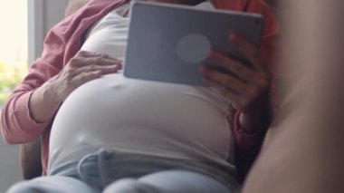 Hamilelik bilgileri için tablet kullanan Asyalı hamile bir kadın. Anne kendini mutlu hissederken pozitif ve huzurlu gülümserken çocuğuyla ilgilenirken oturma odasında kanepede uzanmış ev konseptinde..