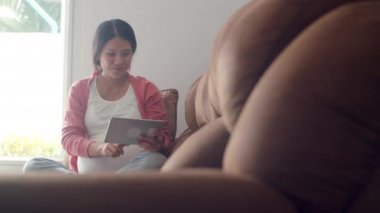 Genç Asyalı Hamile kadın tablet arama gebelik bilgileri kullanarak. Anne mutlu olumlu ve huzurlu gülümseyerek hissederken, evde oturma odasında kanepede yatan çocuğuna dikkat eder. Yavaş çekim.