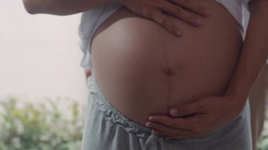 Genç Asya Hamile çift sarılmak ve çocukları ile konuşurken göbek tutarak. Anne ve baba mutlu gülümseyerek huzurlu bir şekilde bakarken bebek, evde oturma odasında pencerenin yanında hamilelik. Yavaş çekim.