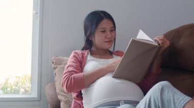Genç Asya Hamile kadın göbek bebek için bir kitap okuyun. Anne mutlu olumlu ve huzurlu gülümseyerek hissederken evde oturma odasında kanepede yatan çocuğa özen derken konsept. Yavaş çekim.